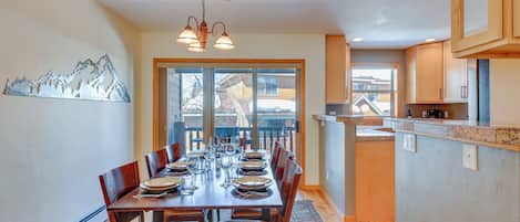 Un comedor con una mesa de madera dispuesta para seis personas, rodeada de sillas, una araña y vistas a través de grandes ventanales.