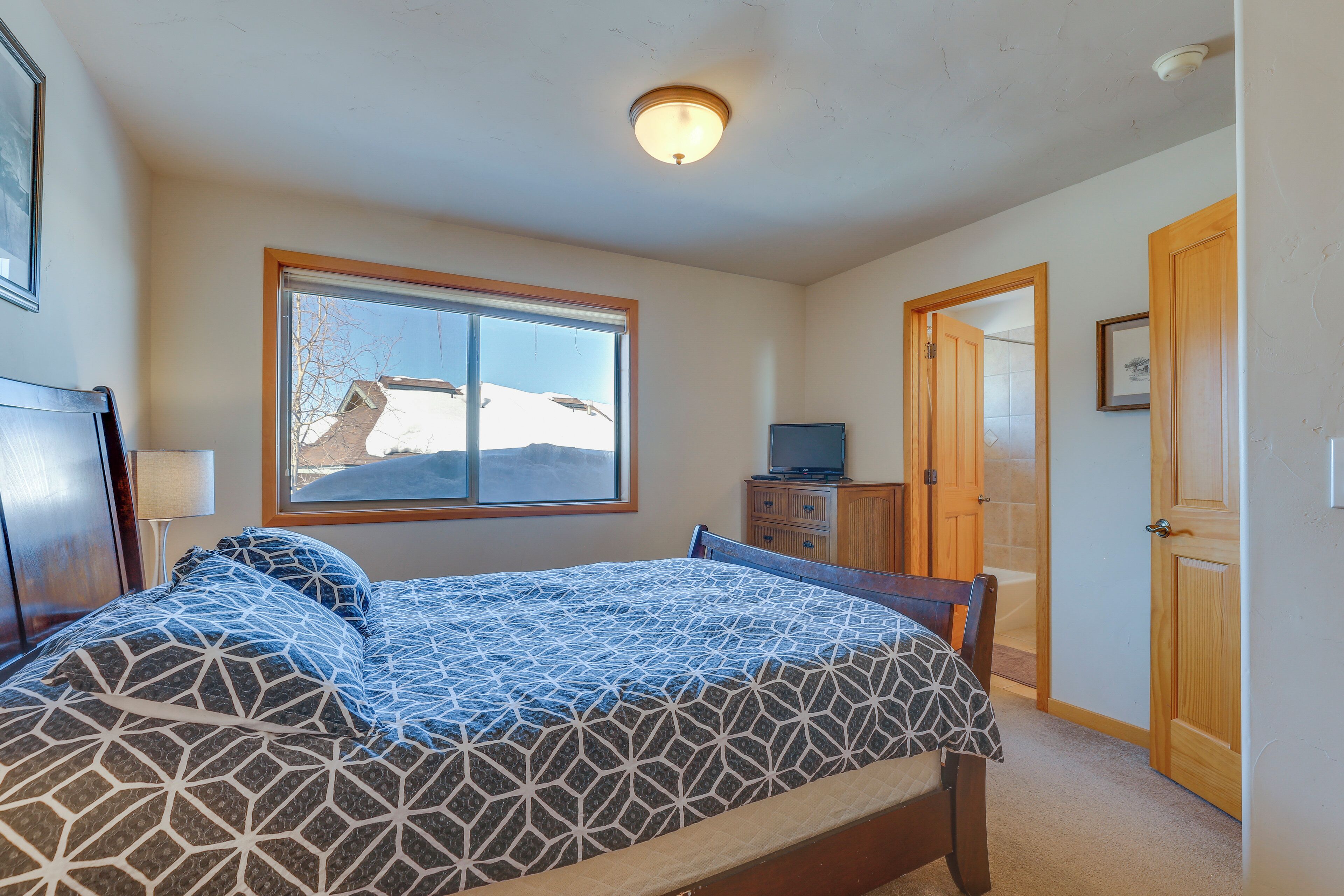 A bedroom with a large bed, a desk with a computer, a window with a view of a snowy landscape, and a door leading to another room.