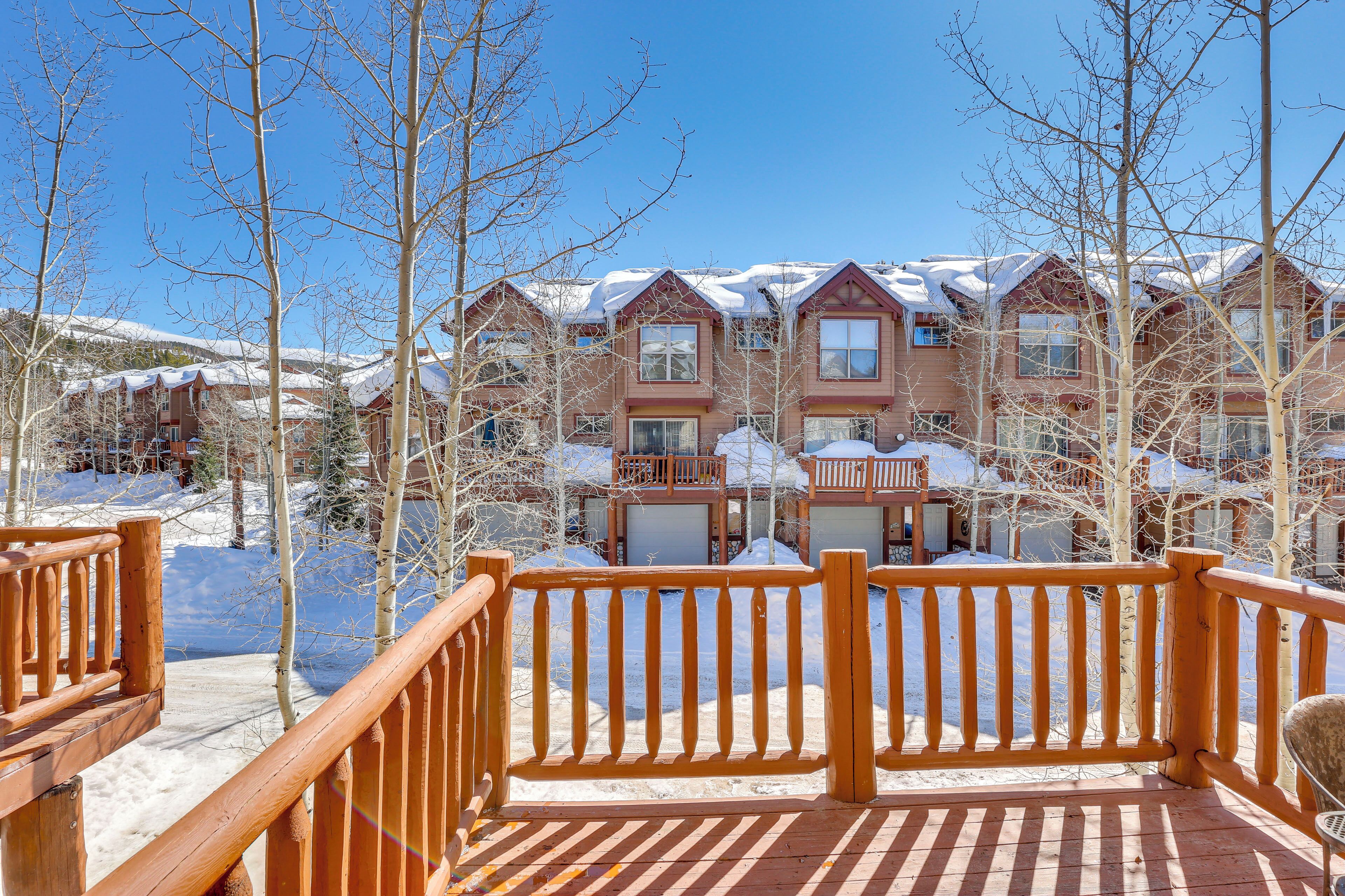 A wooden deck with a railing, snow-covered buildings, and leafless trees in the background.
