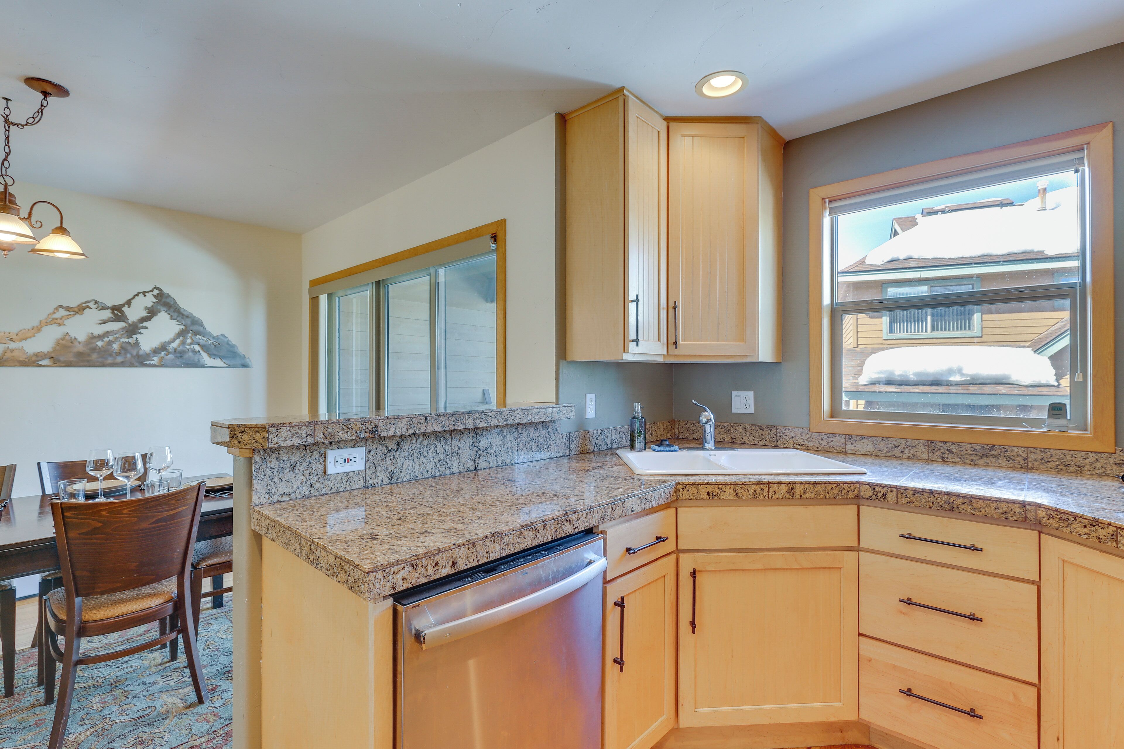 A kitchen with granite countertops, wooden cabinets, a dishwasher, and a window with a view of a house.