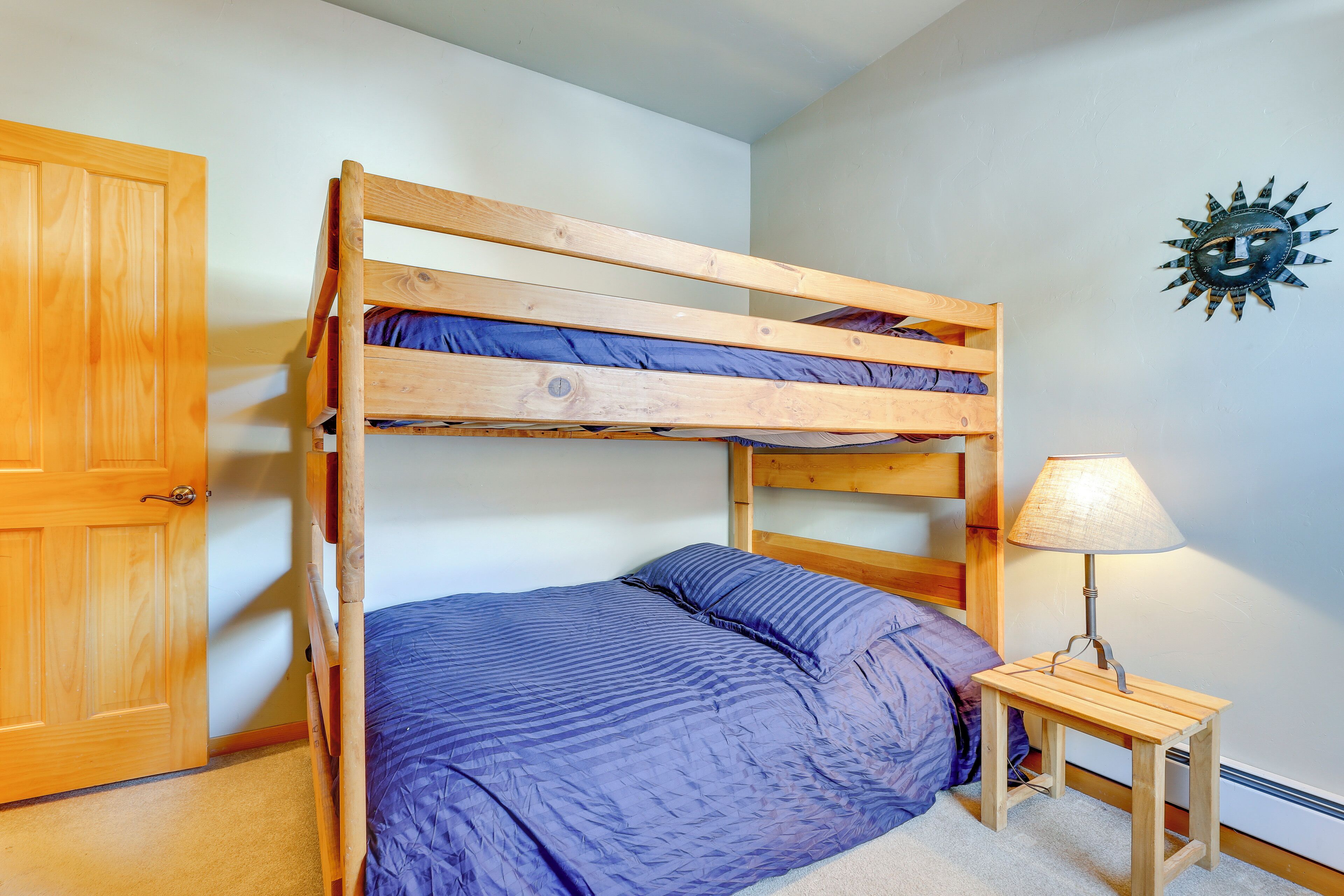 A bunk bed with a blue mattress, a wooden bedside table with a lamp, and a wooden door.