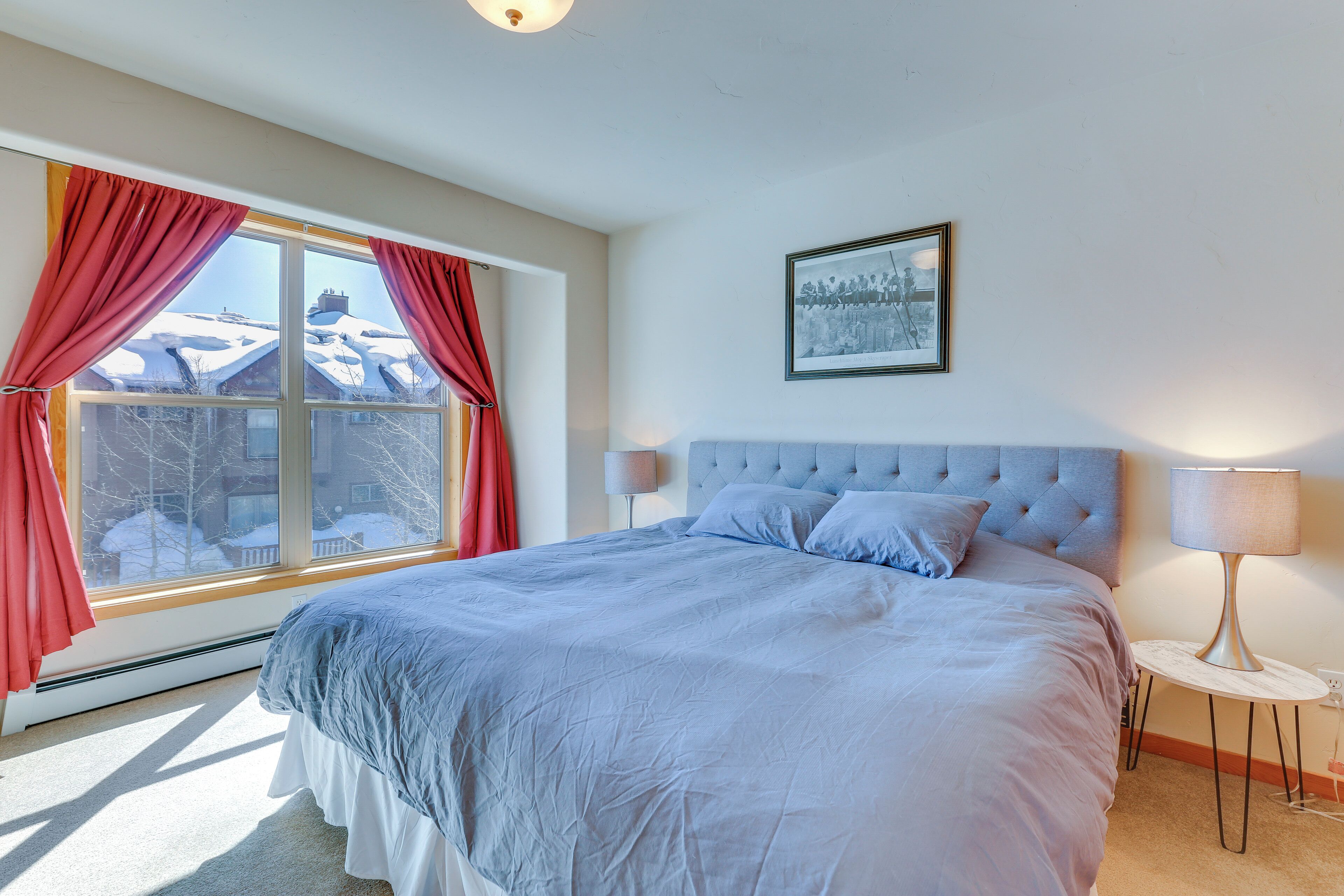 Ein Schlafzimmer mit einem großen Bett, einem Fenster mit roten Vorhängen, einem gerahmten Bild an der Wand und einem Nachttisch mit einer Lampe.