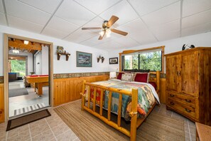 Un dormitorio con cama de madera, ventilador de techo, ventana con vista a árboles, armario de madera y puerta que da a un baño.