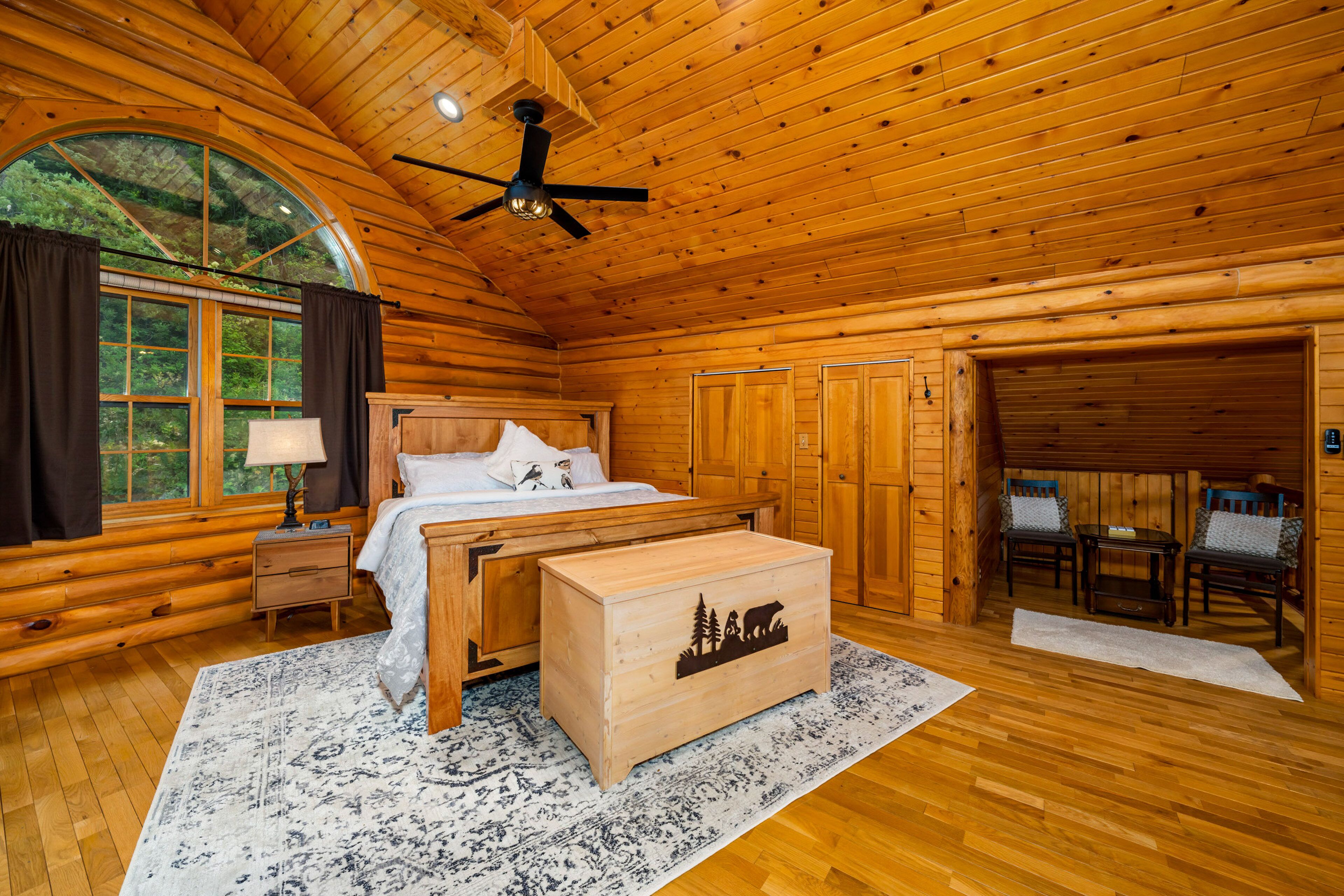 A log cabin bedroom with a bed, nightstand, dresser, and a large window with curtains.