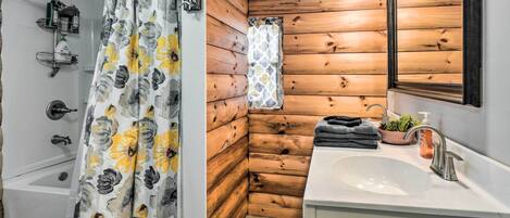 A bathroom with a wooden wall, a shower curtain with a floral pattern, a sink with a mirror, and a small window with a patterned curtain.