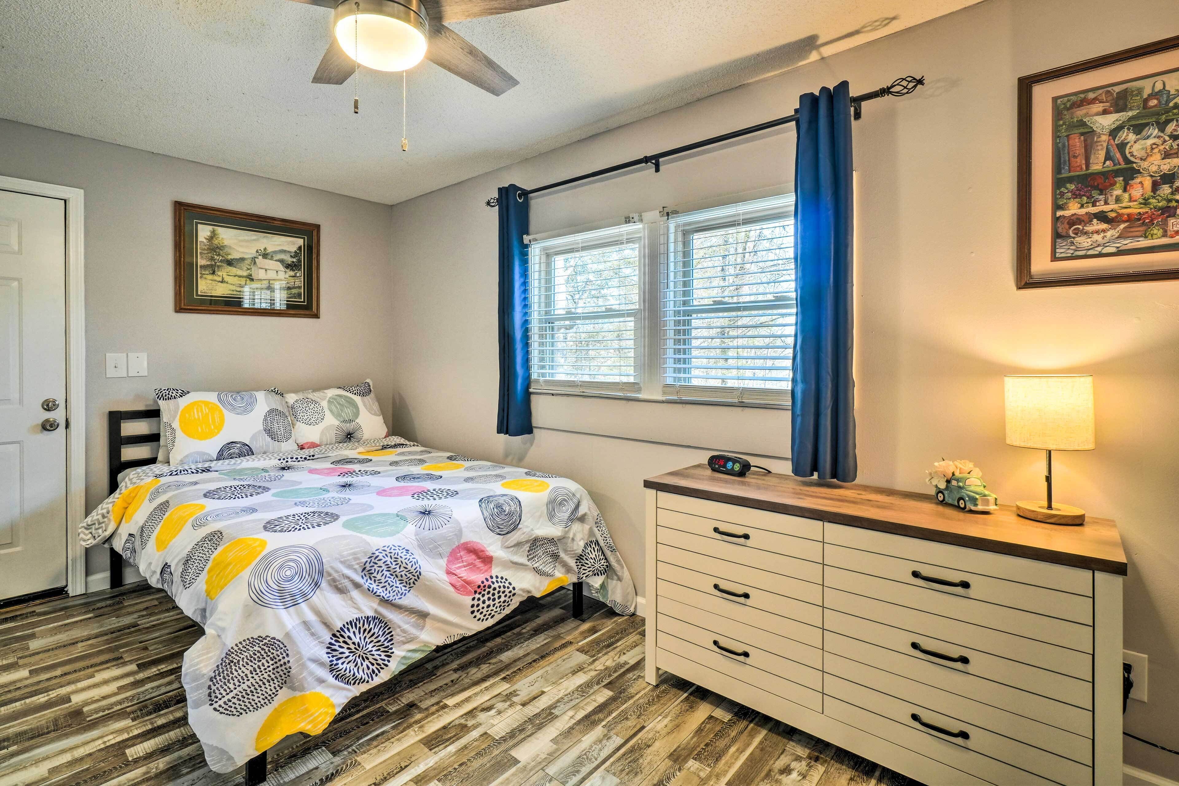 Une chambre à coucher comprenant un lit, une commode, une fenêtre avec des rideaux, un ventilateur de plafond et des œuvres d’art murales.