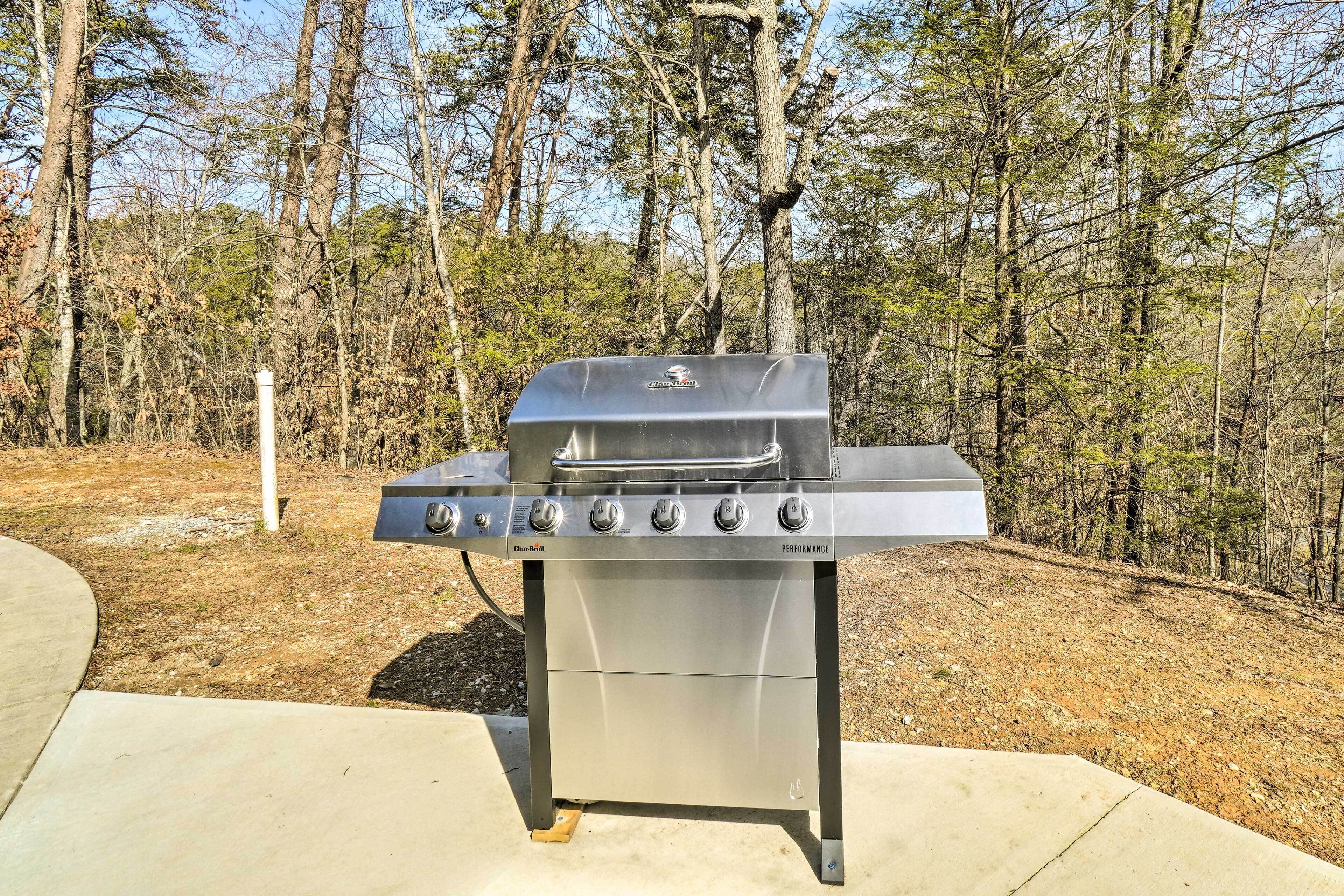 A portable gas grill with four burners and a side shelf, set on a concrete surface in a wooded area.