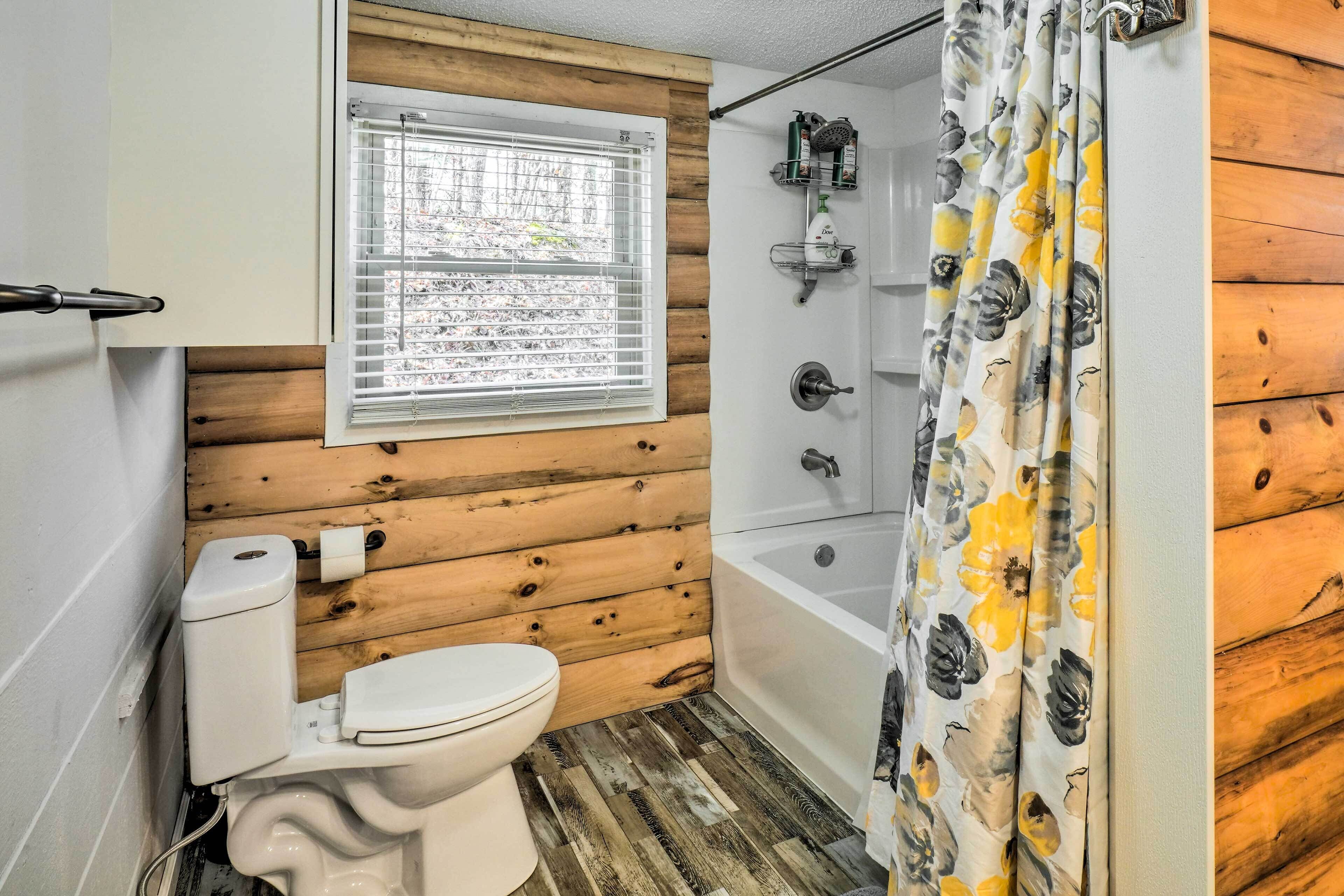 Une salle de bain avec des toilettes, une fenêtre avec des stores, une douche avec un rideau à motifs floraux et un lambris en bois.