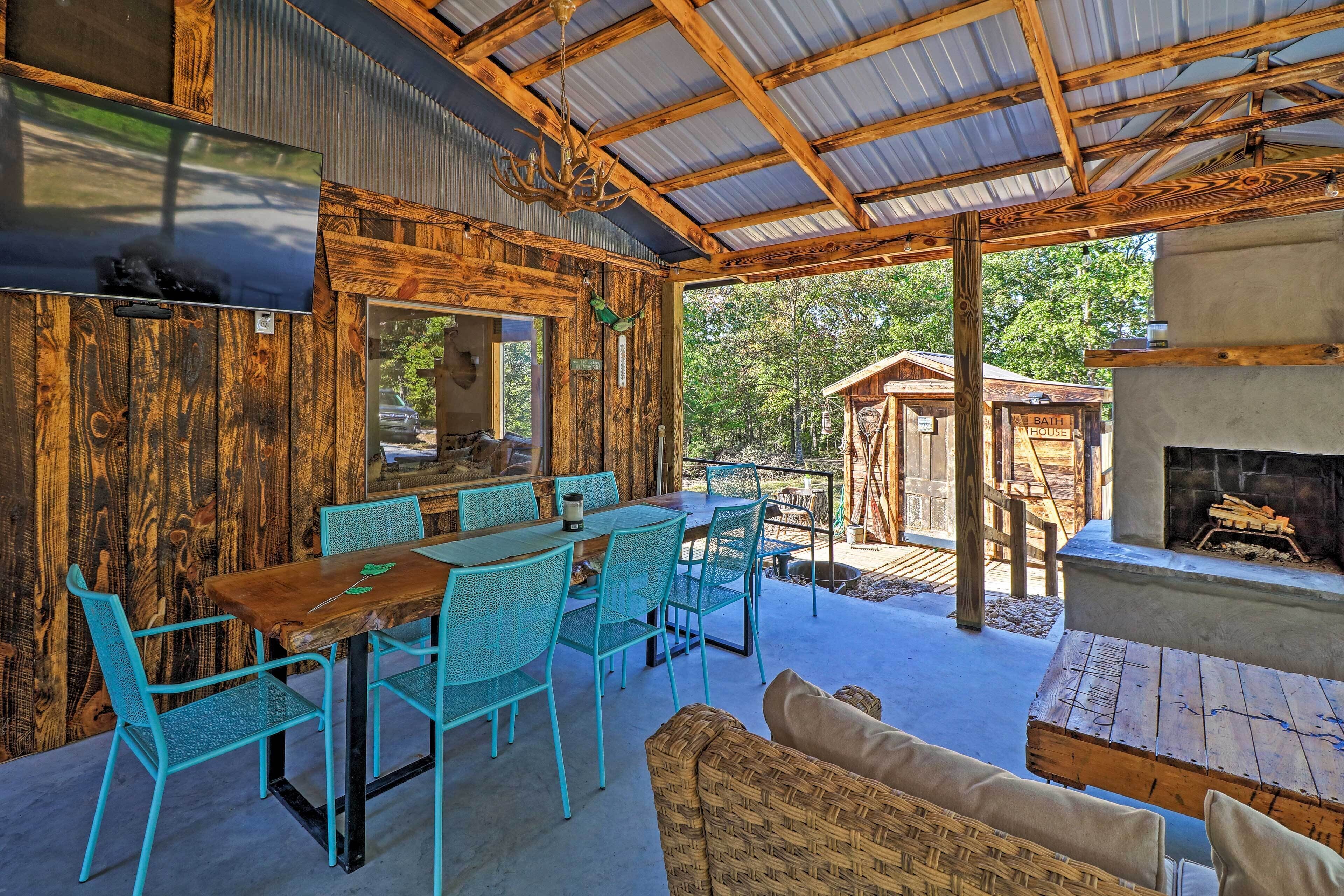 A rustic outdoor dining area with a wooden table and turquoise chairs, a fireplace, and a view of the surrounding forest.