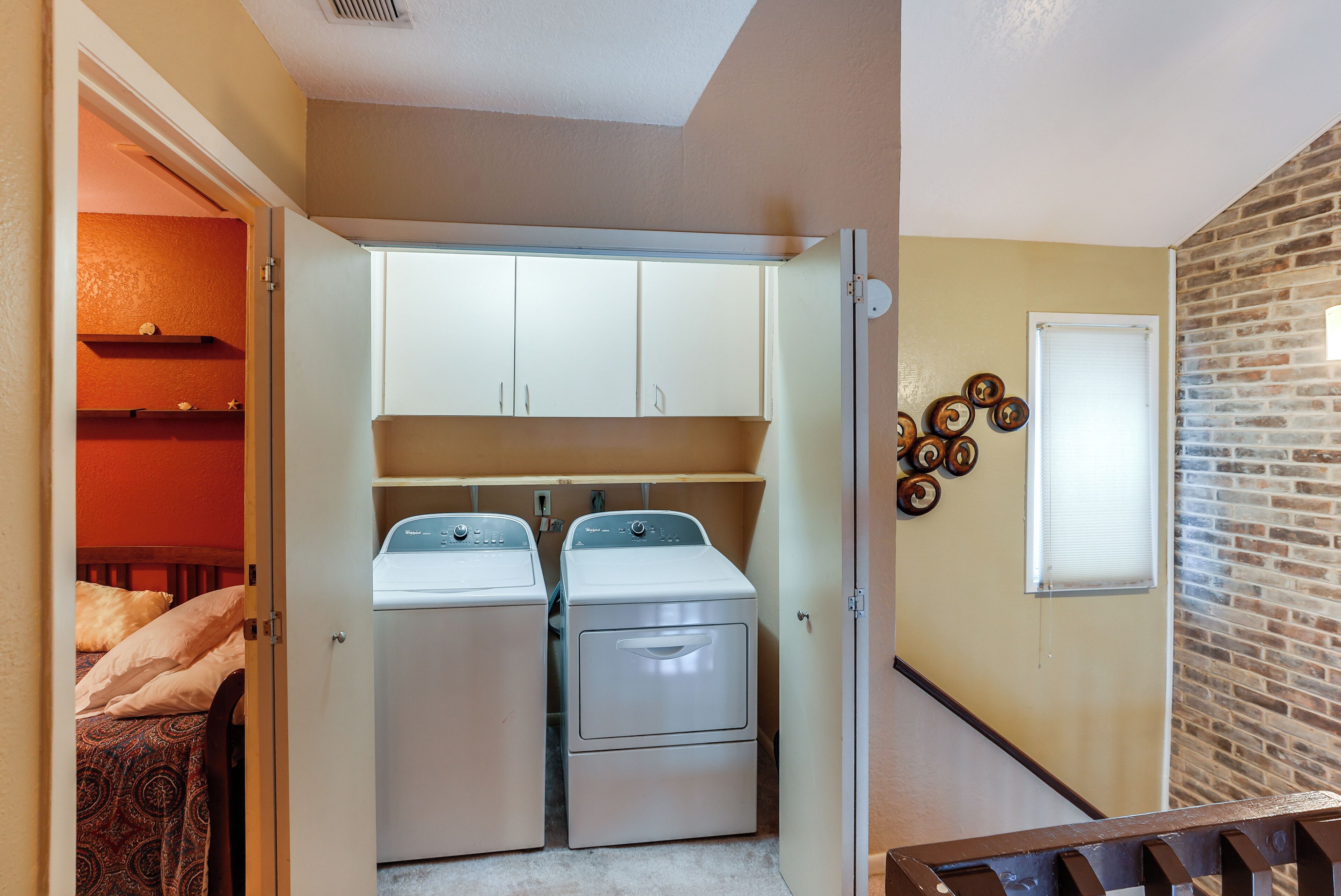 A laundry room with two stacked washers and dryers, a wooden bed with patterned bedding, and a brick wall.