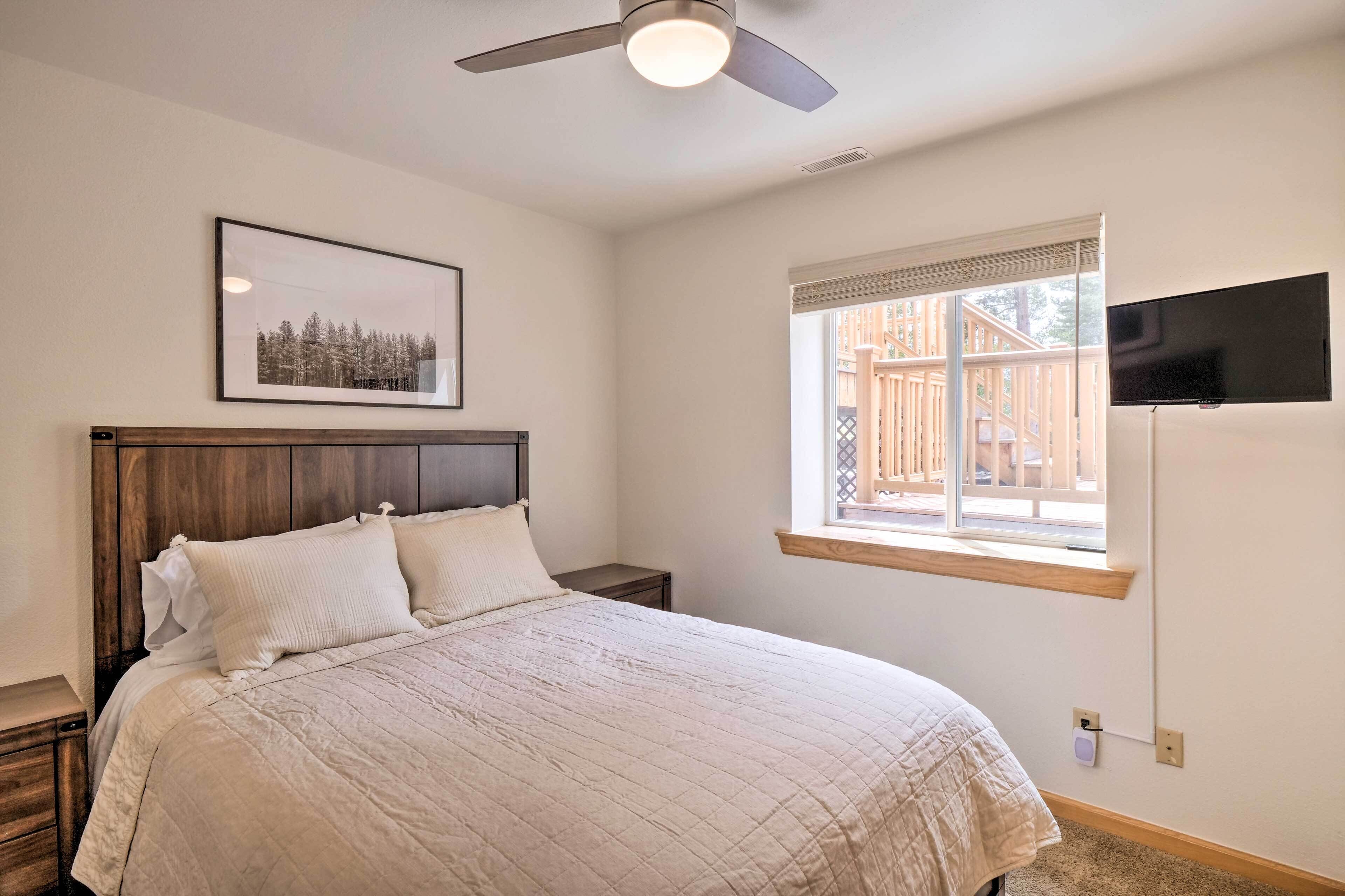 A bedroom with a bed, a ceiling fan, a window with blinds, a mounted TV, and a framed picture on the wall.