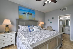 A bedroom with a bed, two nightstands with lamps, a ceiling fan, a painting of boats, decorative wall stickers, and a view into the kitchen. - Sunny SPI Las Villas Condo w/ Resort Amenities! (South Padre Island)