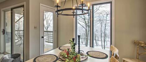 A dining area with a chandelier, round tables, woven placemats, and a view of trees through large windows.