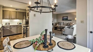 Une cuisine moderne dotée d’appareils électroménagers intégrés, un coin repas avec une table blanche et des sets de table en fibres naturelles, et un salon avec un canapé gris et des coussins décoratifs.
