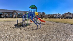 Un parque infantil con un tobogán y estructuras para escalar, rodeado de grava y edificios residenciales al fondo.