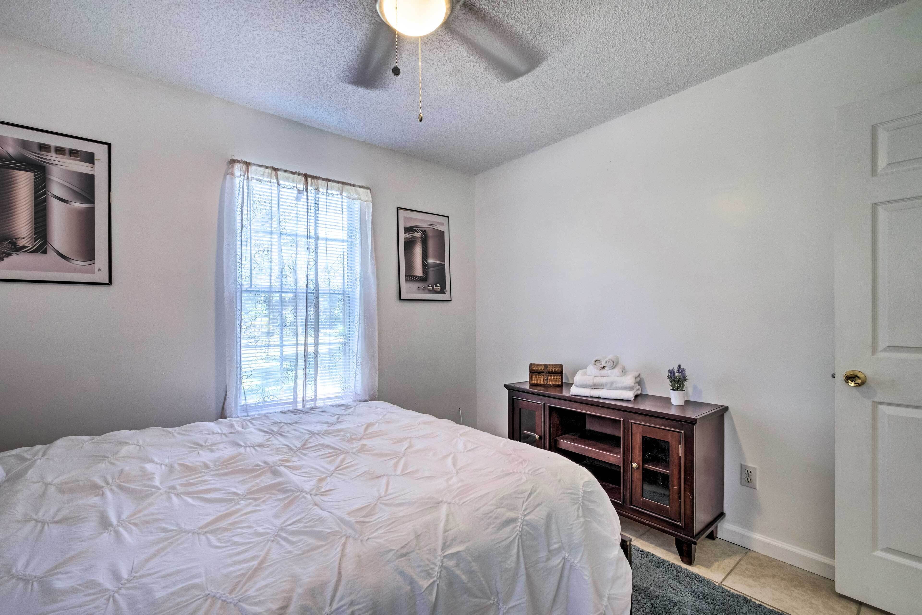 Une chambre à coucher comprenant un lit, un ventilateur de plafond, une fenêtre avec des rideaux, une commode et une porte.