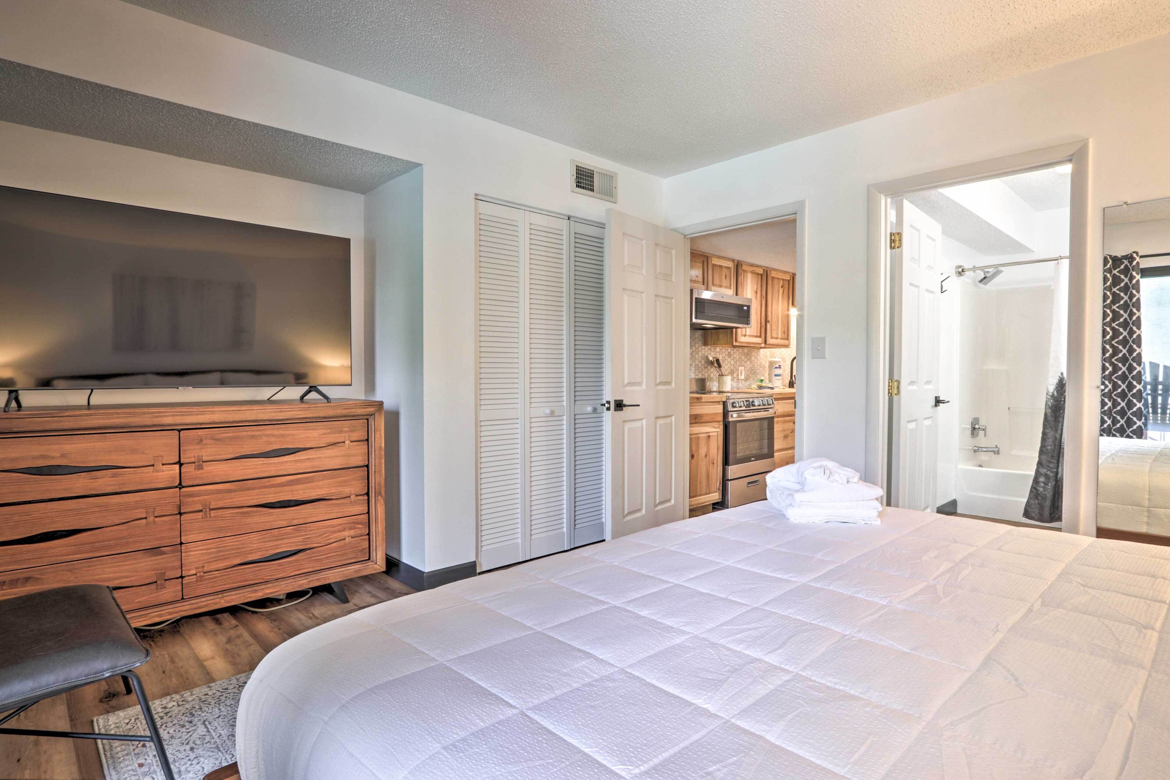 Une chambre à coucher avec un grand lit, une commode en bois, un téléviseur et, au fond, un coin cuisine.