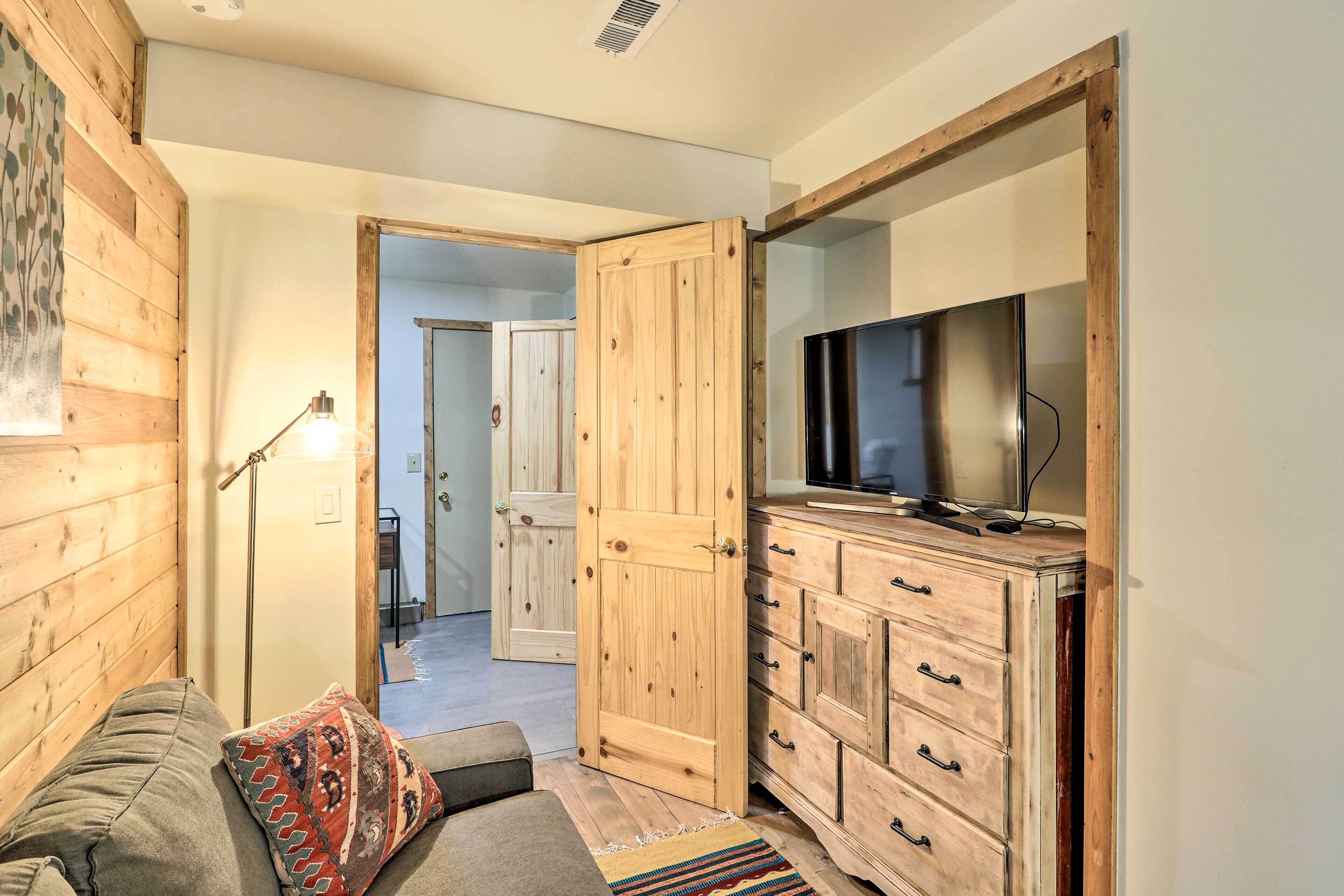Una habitación acogedora con piso de madera, un sofá, un televisor sobre un mueble de madera y una puerta que da a otra habitación.