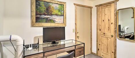 Une pièce avec un bureau en verre, une chaise, un téléviseur, un tableau, une porte en bois et un miroir.
