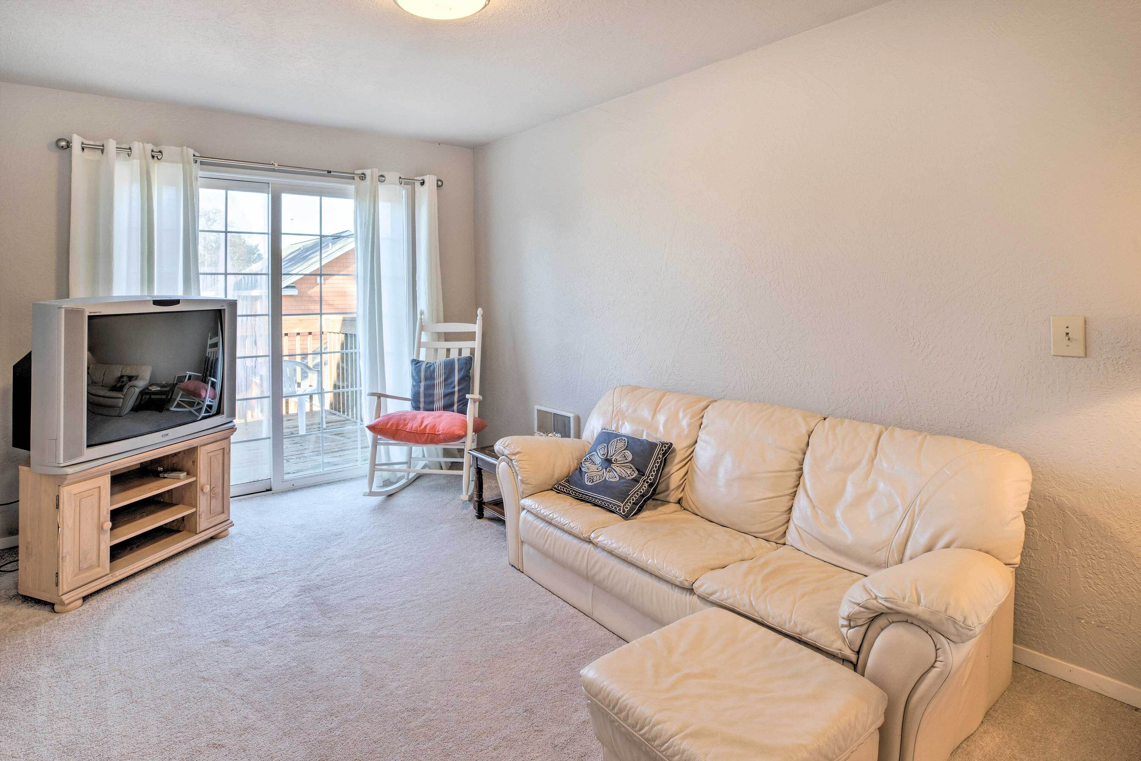 Een woonkamer met een beige leren bank, een televisie op een houten standaard, een schommelstoel en een glazen schuifdeur naar een balkon.