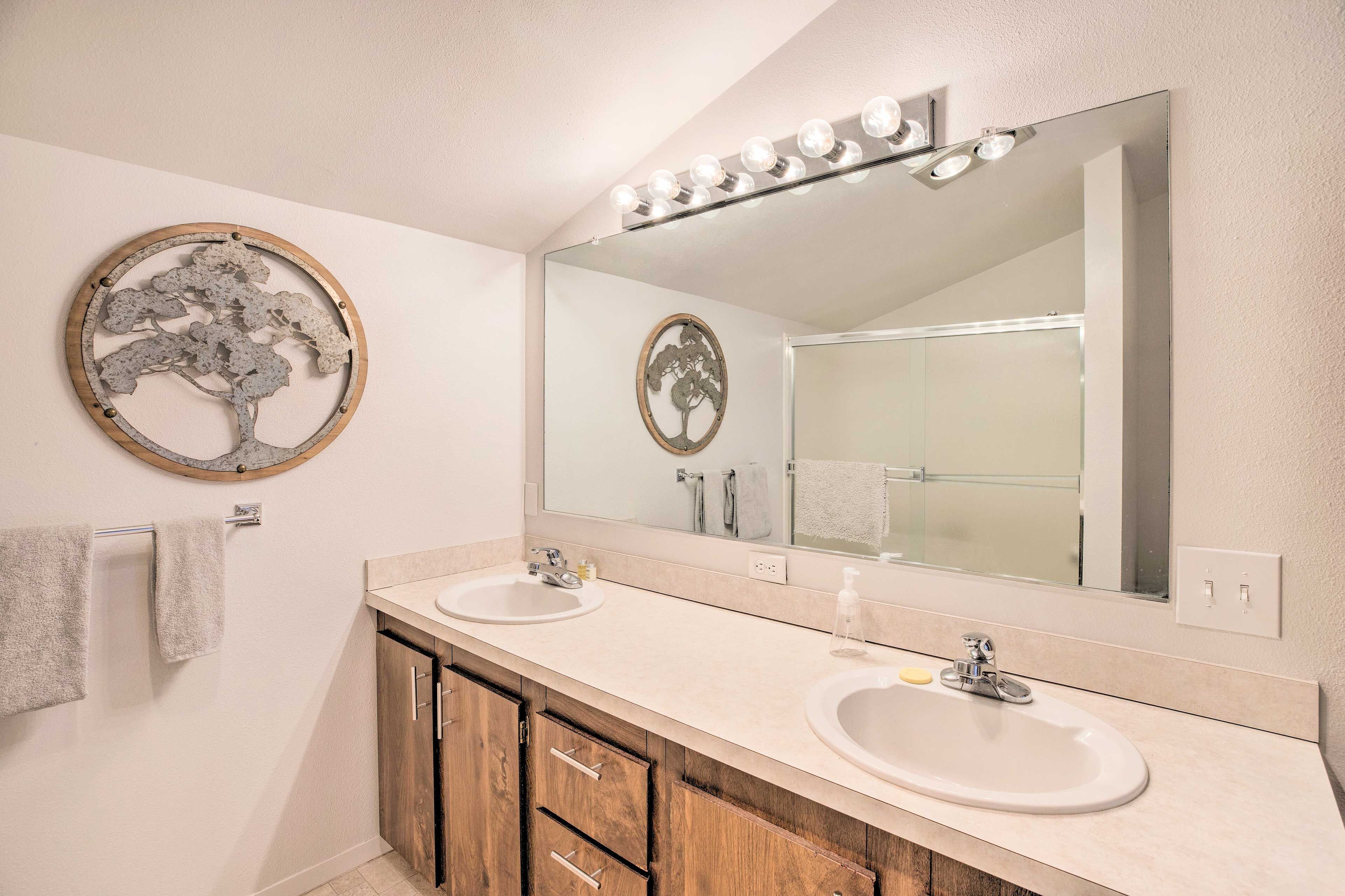 A bathroom with two sinks, a large mirror with lights, and a decorative tree wall art piece.