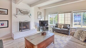 A living room with a fireplace, a television, a sofa, and large windows. - Pet-friendly Rockaway Beach Home w/ Fire Pit! (Rockaway Beach)