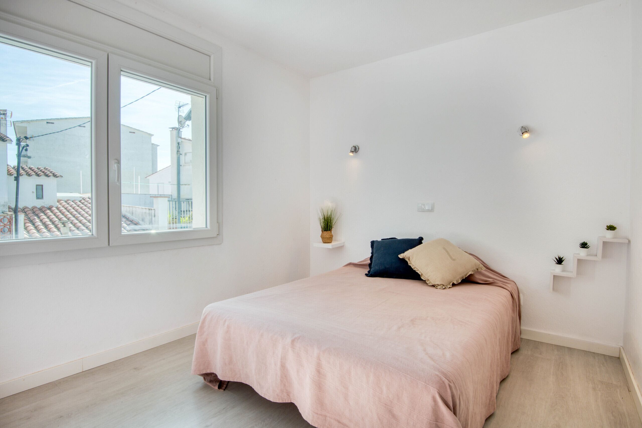 Une chambre avec une grande fenêtre, un lit avec une literie rose et de petites plantes accrochées au mur.