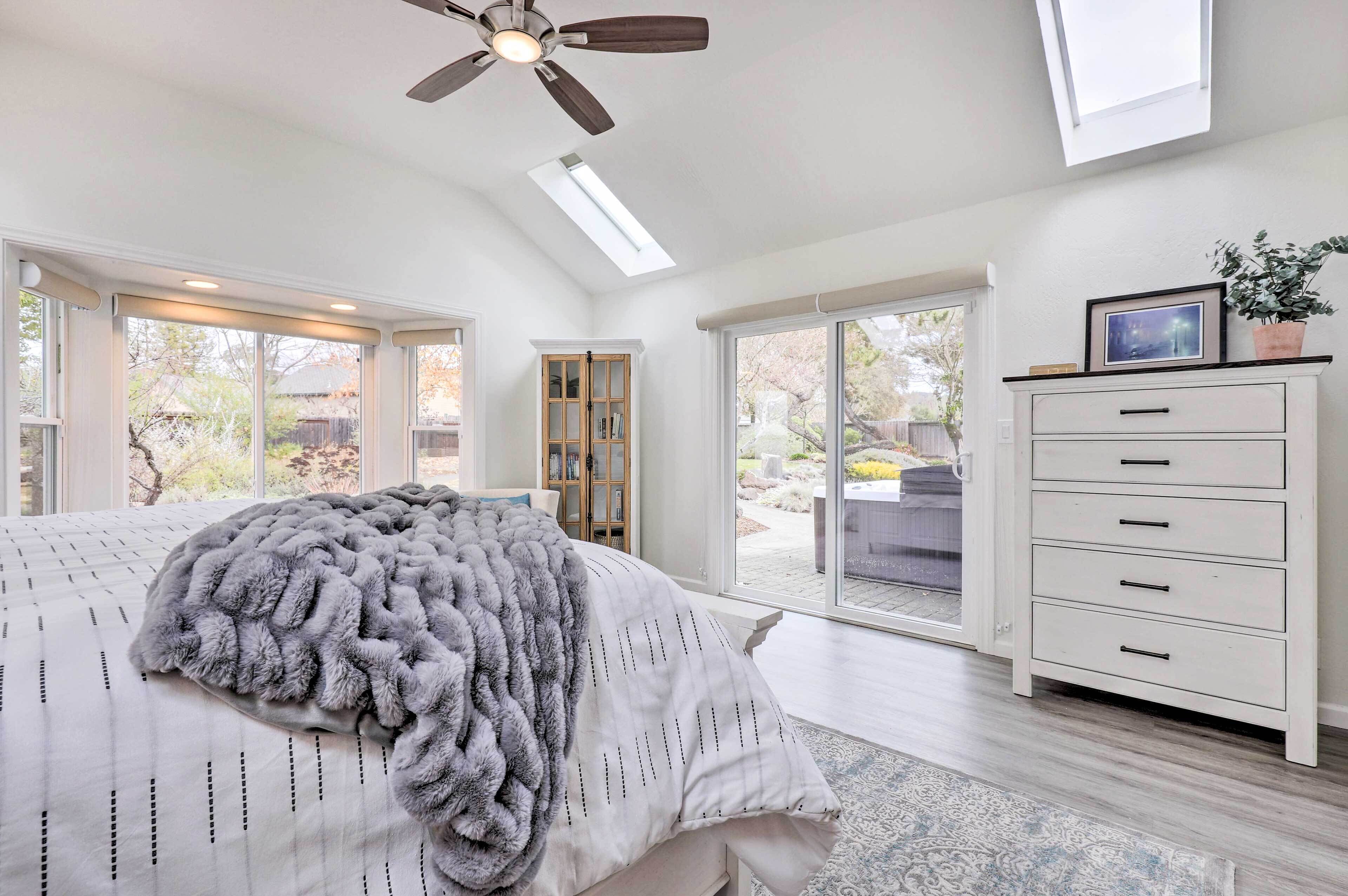 A bedroom with a large bed, a ceiling fan, a skylight, a white dresser, and a view of the outdoors.