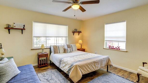 A bedroom with a bed, two windows with blinds, a ceiling fan, a bedside table with a lamp, and a small shelf with decorative items. - Charming Home w/ Yard ~ 5 Mi to Clearwater Beach! (Clearwater)