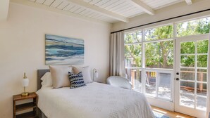 A bedroom with a large bed, a nightstand with a lamp, a painting on the wall, and a view of the outdoors through a sliding glass door. - Vacation Rental Home ~ 1 Mi to Carmel Beach! (Carmel)