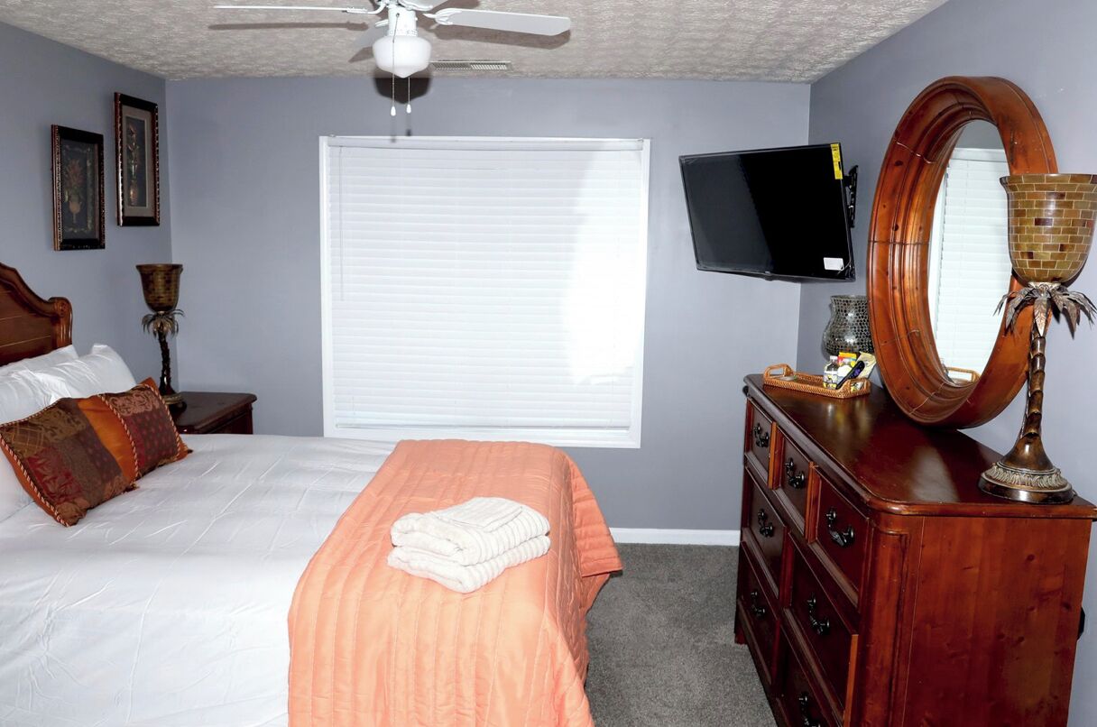 Een slaapkamer met een bed, een dressoir, een televisie, een plafondventilator en een raam met jaloezieën.