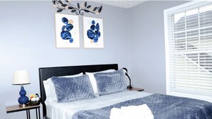 Un dormitorio con cama, mesita de noche, lámpara y una obra de arte en la pared.