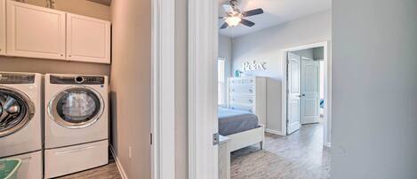A laundry room with a washing machine and dryer, a ceiling fan, and a hallway leading to a bedroom with a bed and dresser.