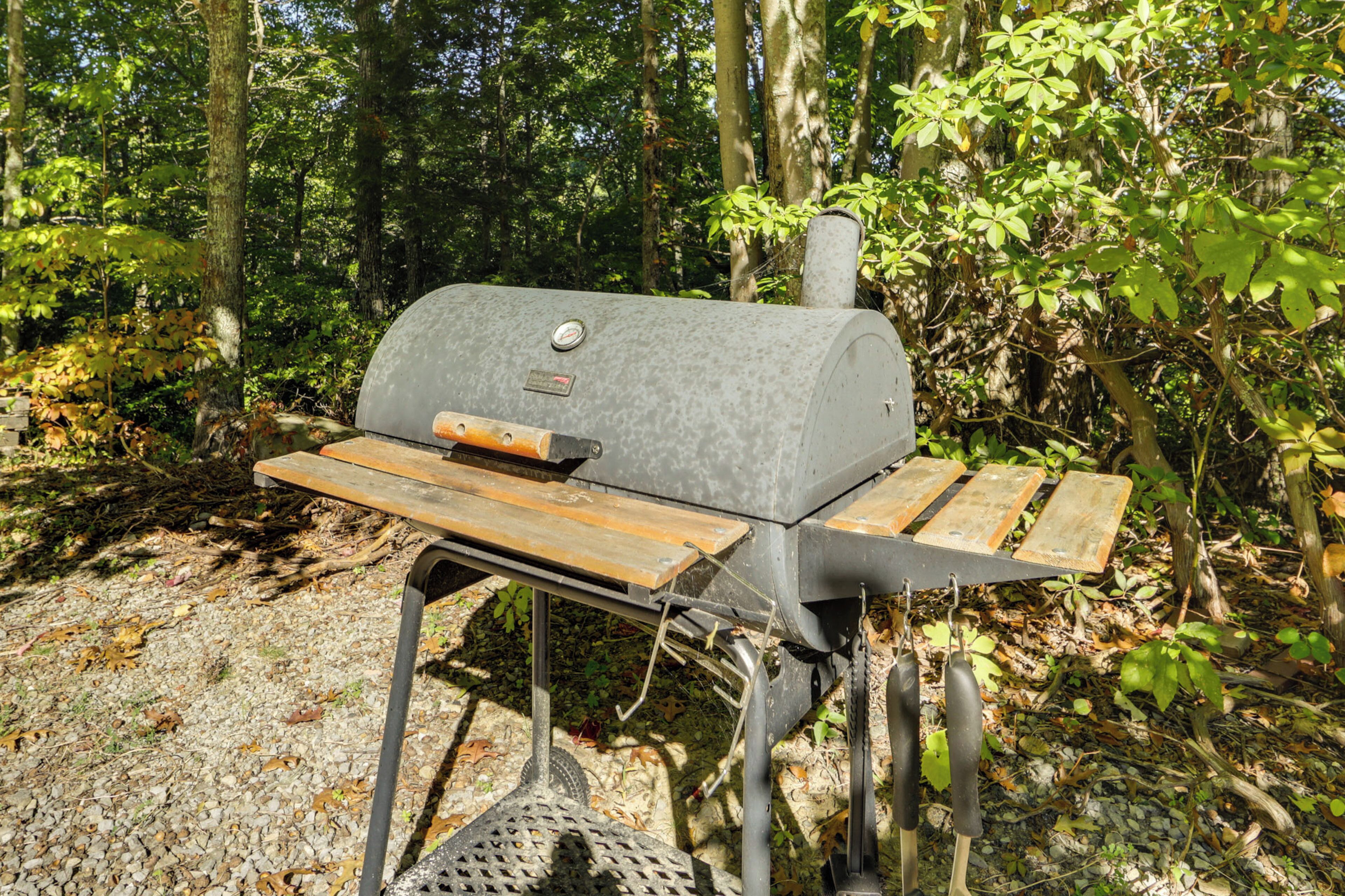 En portabel grill med ett metallställ och träribbor, placerad i ett skogsområde med träd och lövverk.