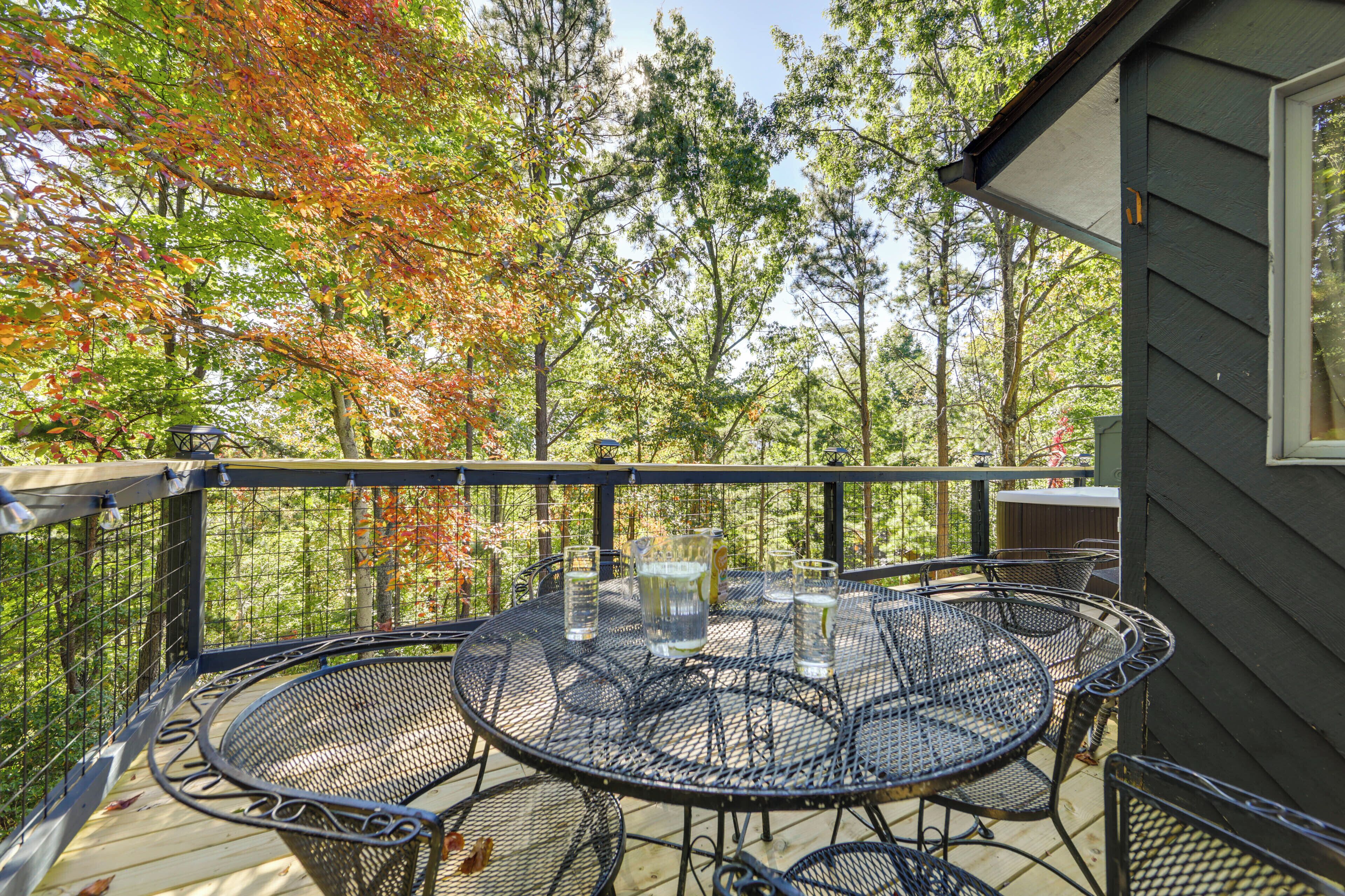 A round metal table with chairs on a wooden deck, surrounded by trees with autumn foliage.