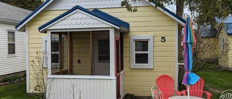 A yellow house with a blue roof, a porch with white railings, and a brick patio with white metal chairs and a colorful umbrella.