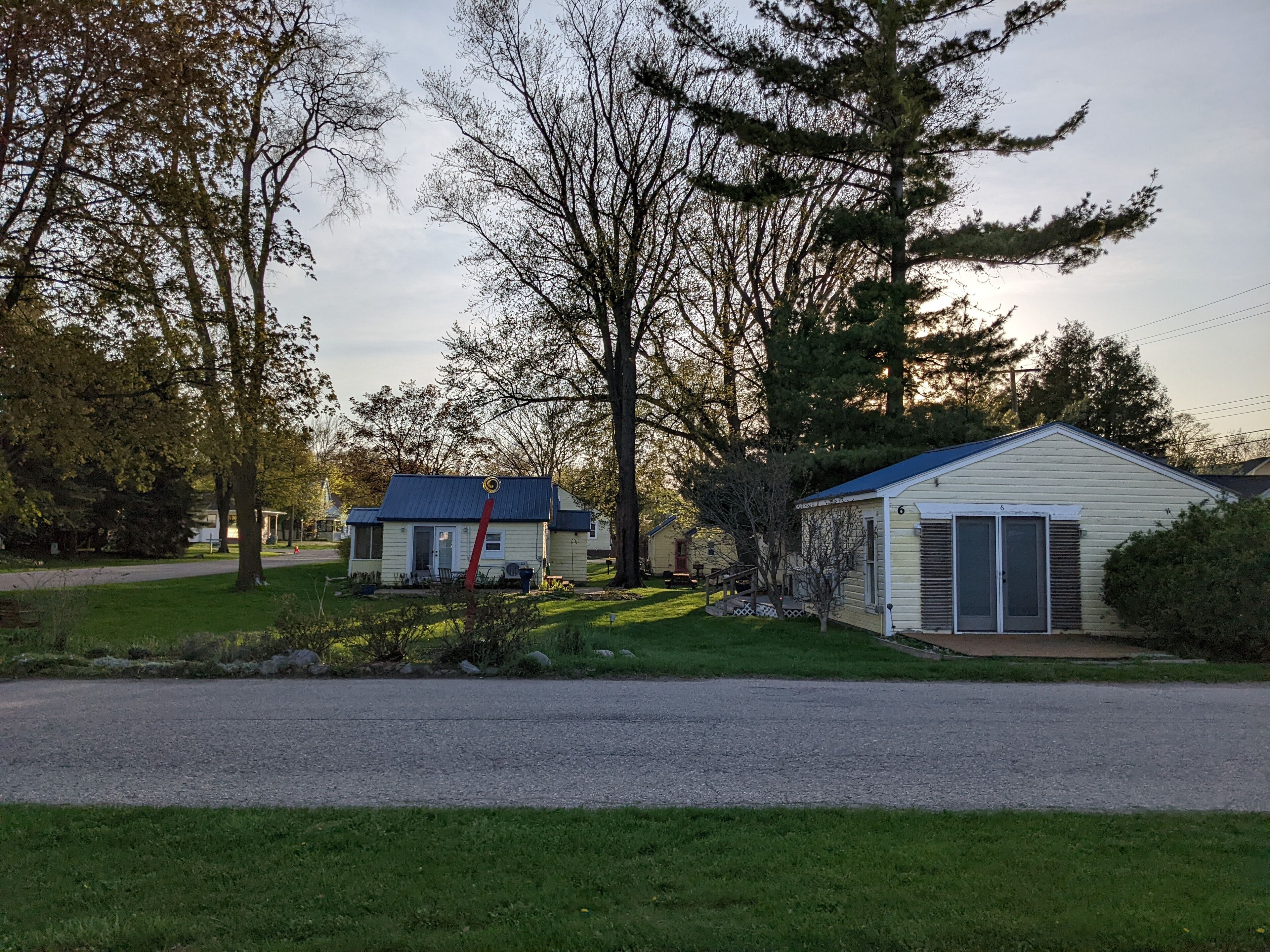 A small, single-story house with a blue roof, surrounded by trees and a gravel driveway.