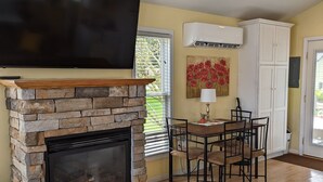 A living room with a stone fireplace, a flat-screen TV mounted above it, a dining area with a table and chairs, and a colorful rug on the wooden floor. - Little Yellow Cottages (Port Austin)