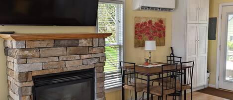A living room with a stone fireplace, a flat-screen TV mounted above it, a dining area with a table and chairs, and a colorful rug on the wooden floor.