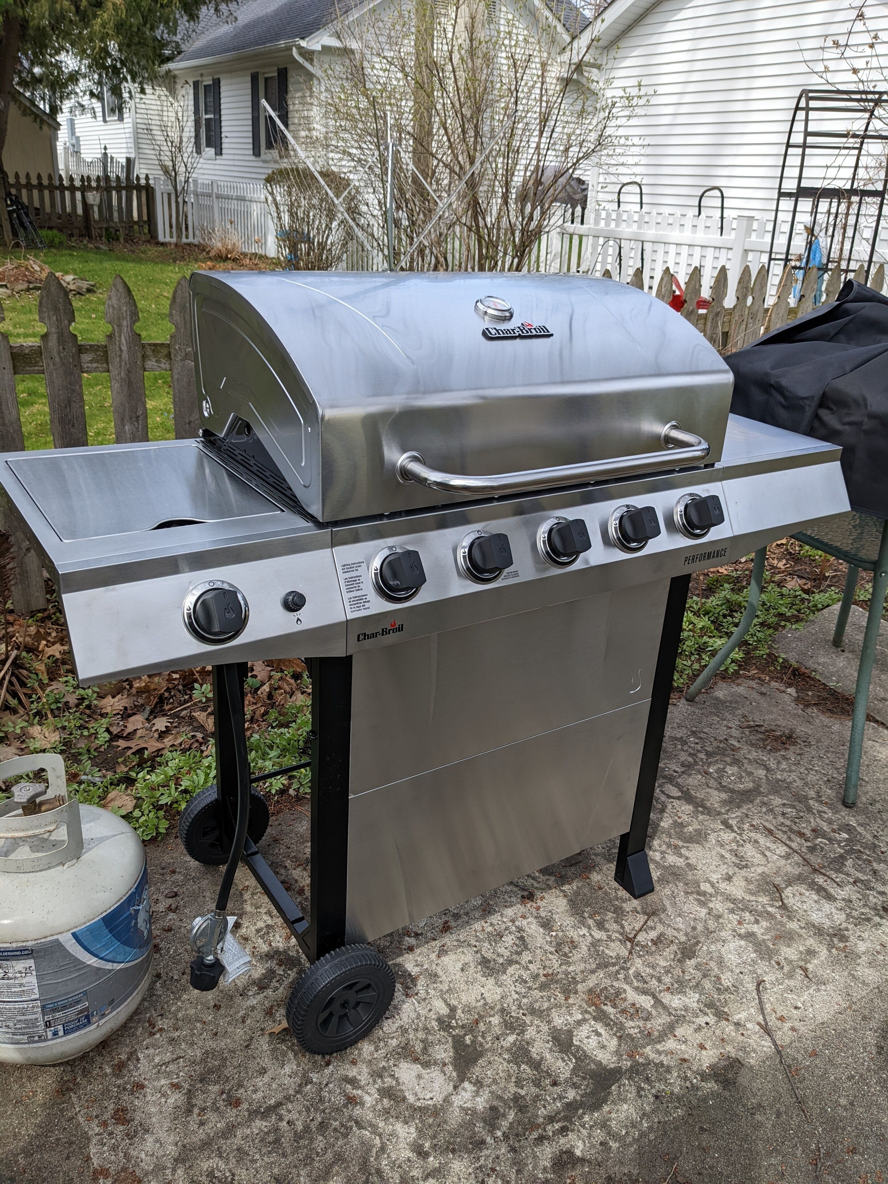 A Char-Broil gas grill with four burners and a side shelf, positioned on a concrete patio with a white fence and house in the background.