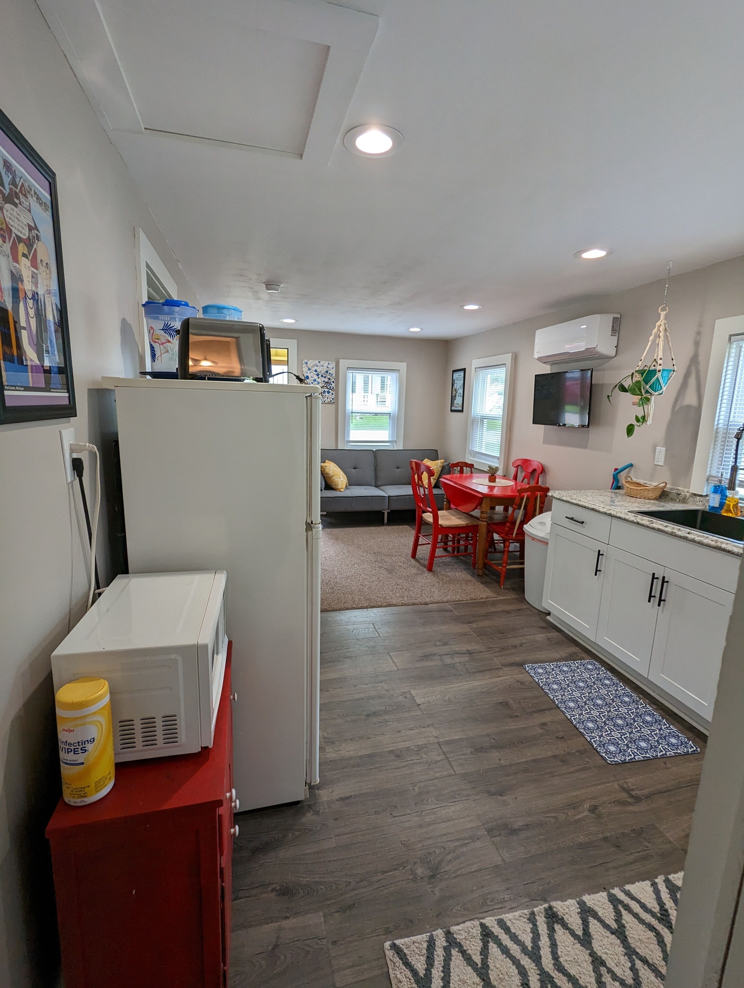 A compact kitchen with a refrigerator, microwave, and a red cabinet with a can of wipes. There is a dining area with a red table and chairs, and a gray sofa.