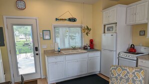 A compact kitchen with white cabinets, a microwave, a stove, and a sink. There is a black rug on the floor, a chair, and a dining table with chairs. - Little Yellow Cottages (Port Austin)