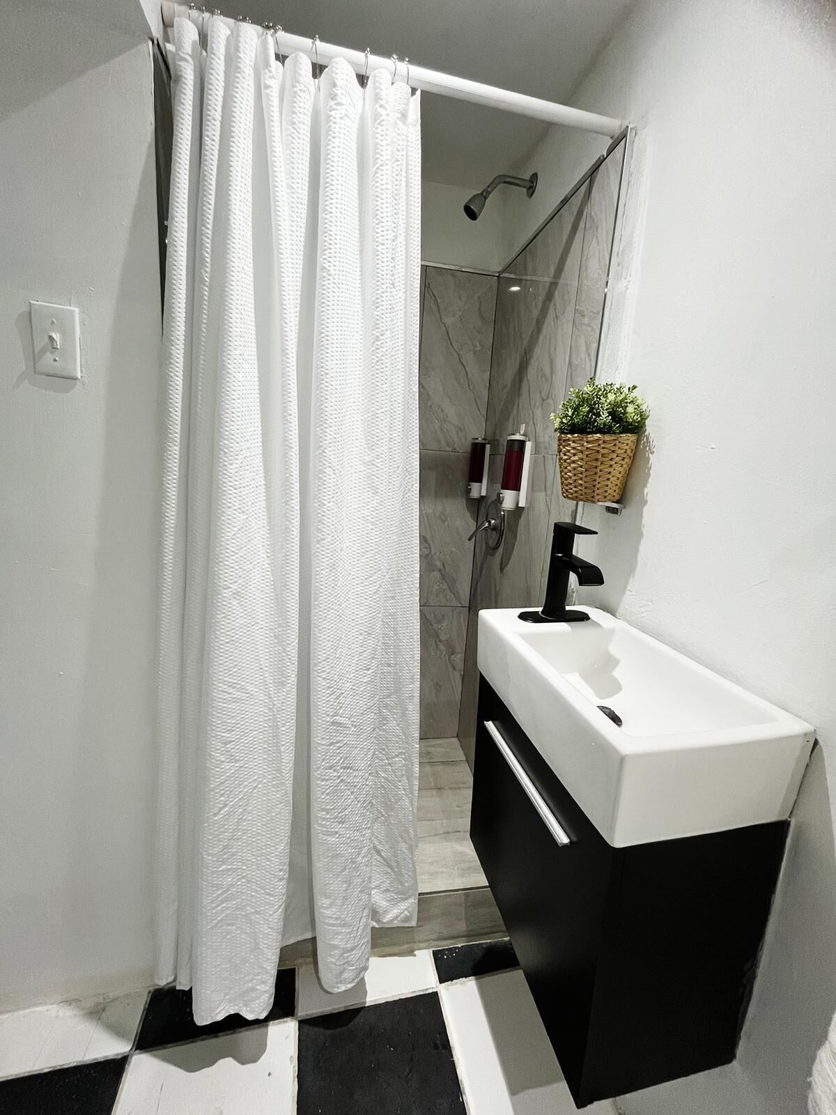 Un baño moderno con cortina de ducha blanca, piso a cuadros blanco y negro, lavamanos blanco con grifo negro, y una pequeña planta en maceta en la pared.