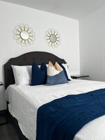 Una cama bien hecha con un cabecero oscuro, ropa de cama blanca y azul, y una manta azul. Hay dos espejos decorativos con marcos dorados fijados a la pared.