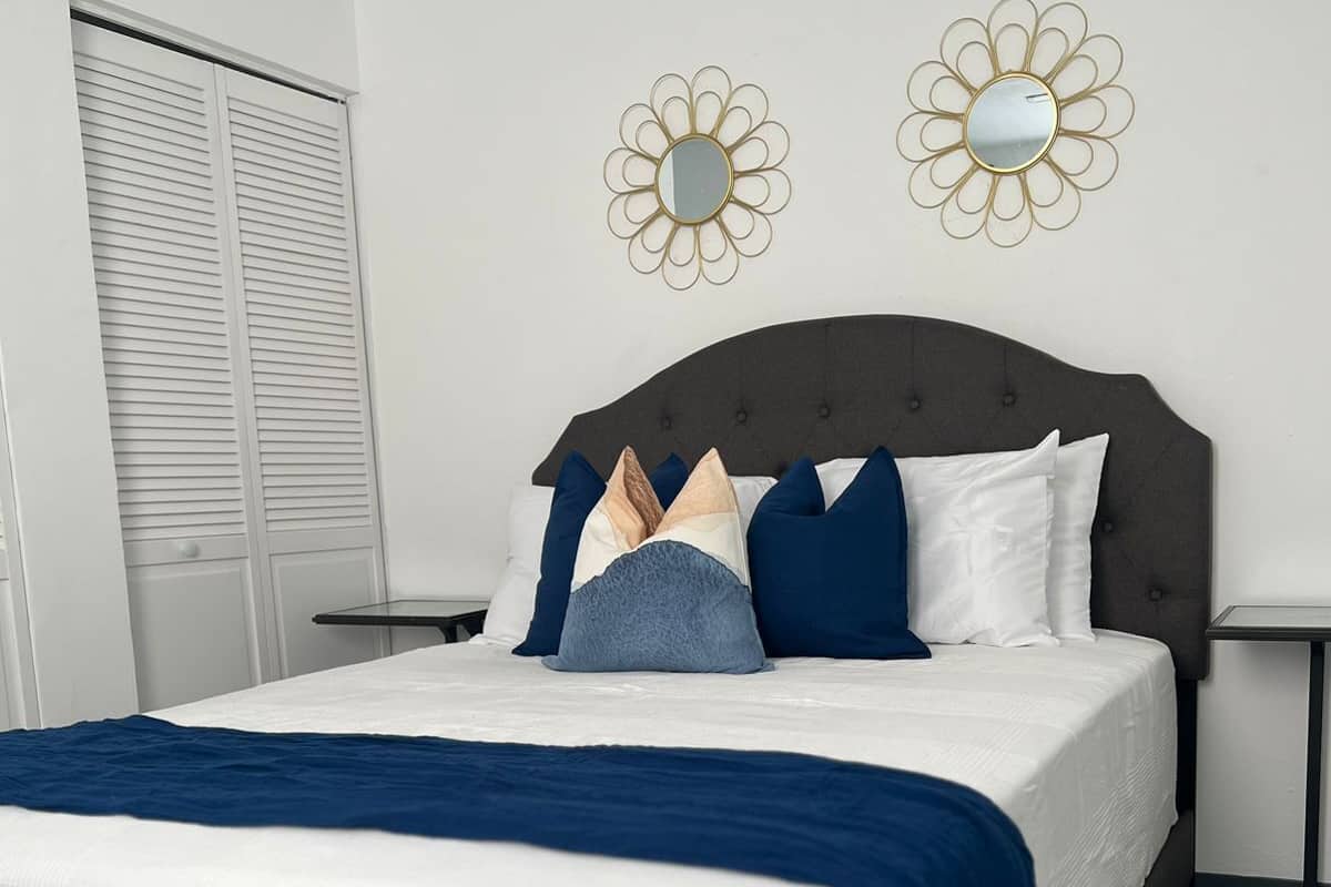 A neatly made bed with a dark headboard, white bedding, and a blue throw blanket. There are two bedside tables with lamps, and a window with closed blinds.