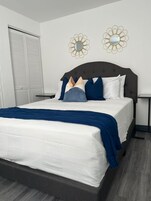 Una cama bien hecha con un cabecero oscuro, ropa de cama blanca y una manta azul. Hay dos mesitas de noche con lámparas, y una ventana con persianas cerradas.