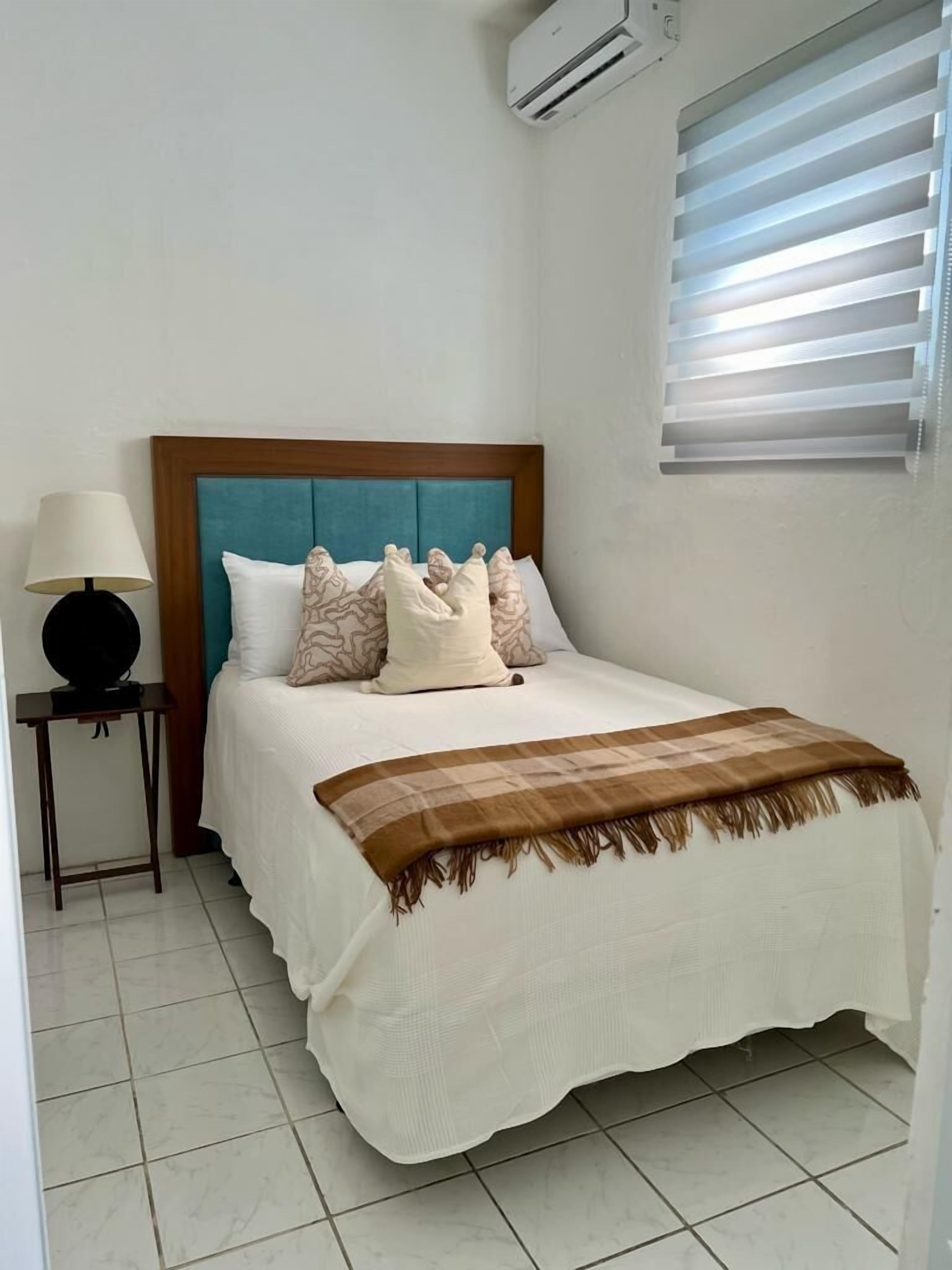 A neatly made bed with a blue headboard, a bedside lamp, and a window with blinds.