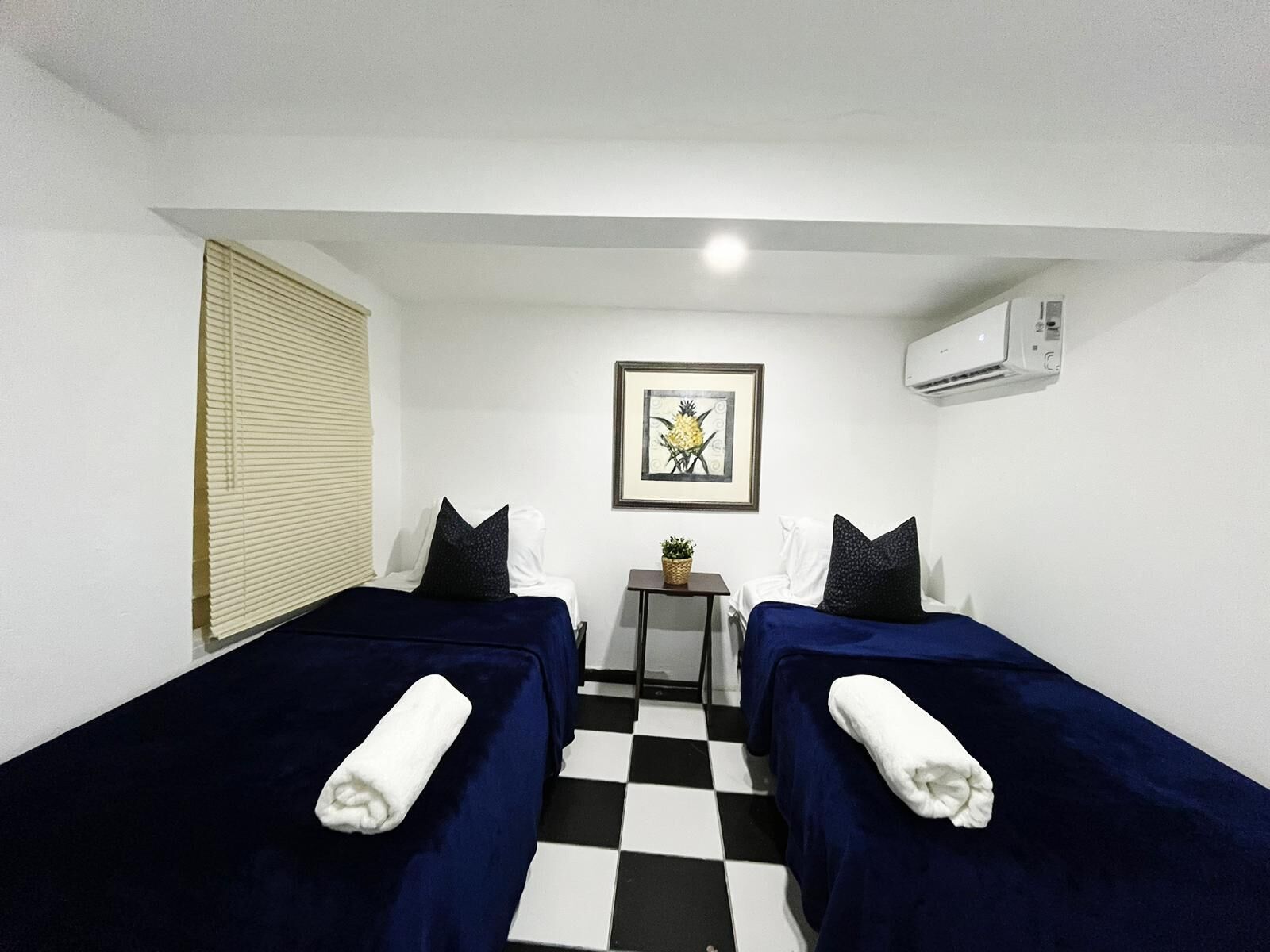 A hotel room with two single beds, a checkered floor, a small table with a plant, and a framed picture on the wall.