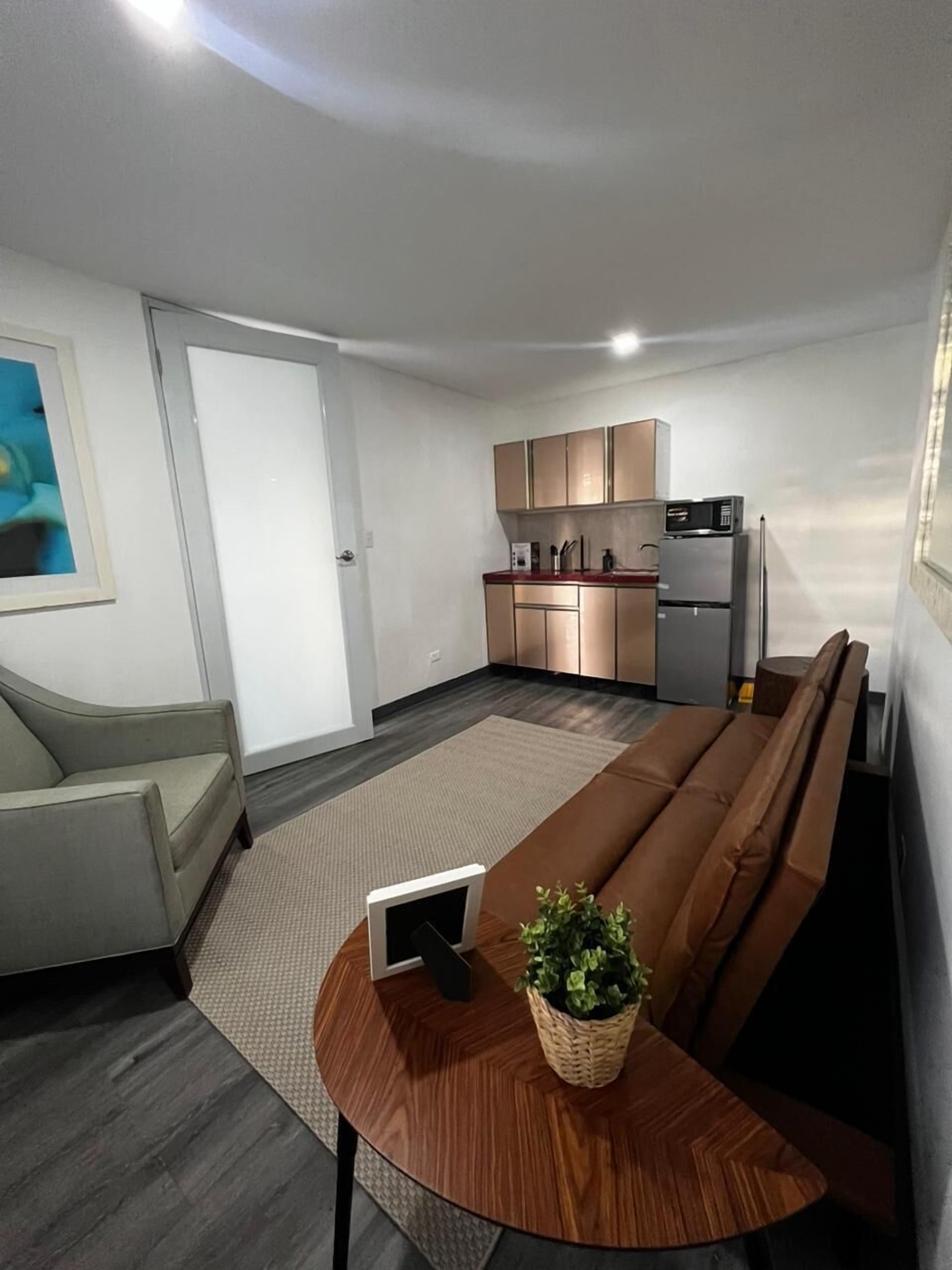 A modern living room with a brown sofa, a small wooden table with a tablet and a plant, a grey armchair, and a kitchen area with cabinets and appliances.