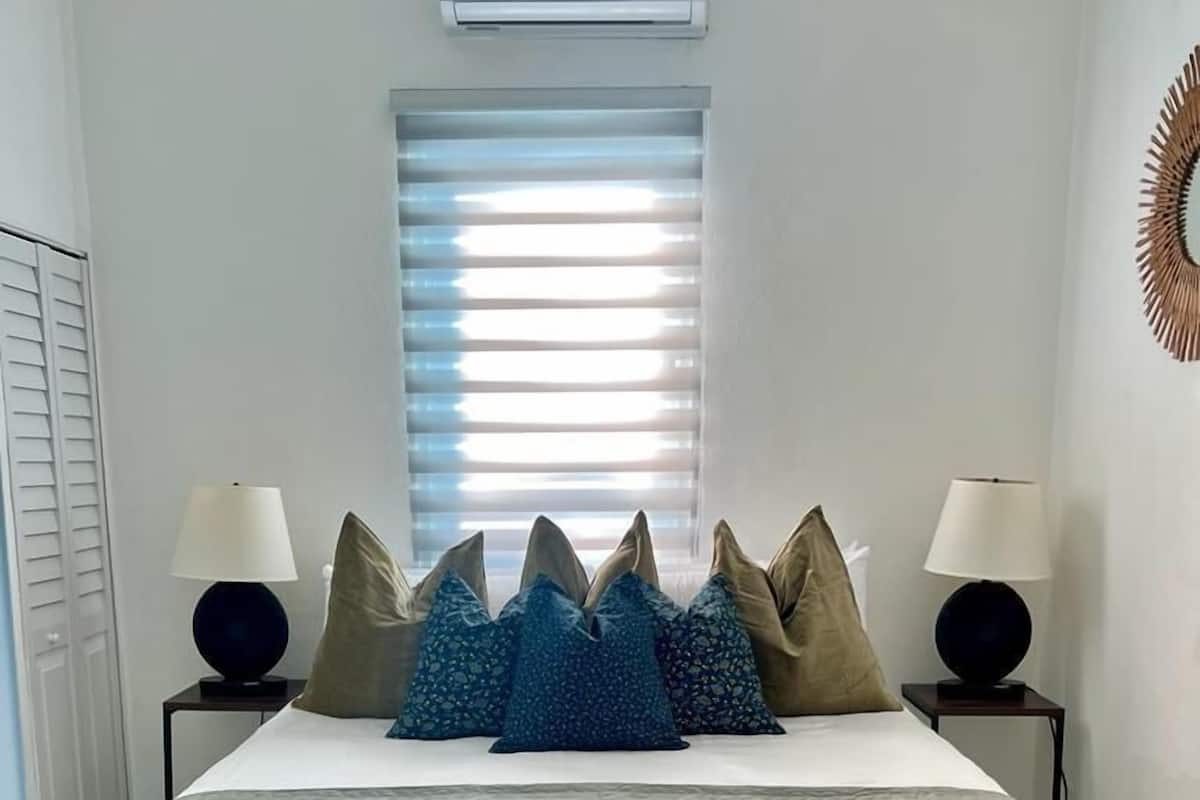 A bedroom with a bed, two bedside tables, a window with blinds, and a wall-mounted air conditioner.